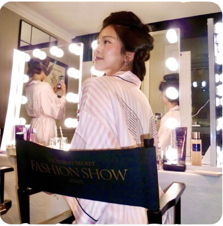 Person in a robe standing in front of a mirror with lights, wearing a 'Victoria's Secret Fashion Show' chair cover.