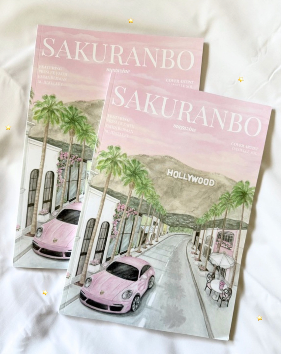 Two copies of the Sakuranbo magazine featuring a pastel pink and white illustration of a street scene with palm trees, mountains, and a pink car, displayed on a white fabric background.